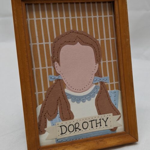 Mid brown framed portrait, paper and stiched simple portrait of Dorothy Gale. At the bottom of the portrait there is a tattoo ribbon banner that says Dorothy