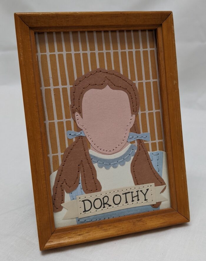 Mid brown framed portrait, paper and stiched simple portrait of Dorothy Gale. At the bottom of the portrait there is a tattoo ribbon banner that says Dorothy